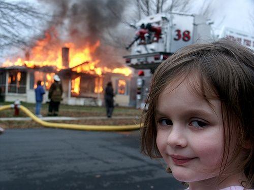 A girl smiles at the camera while a house burns behind her.
